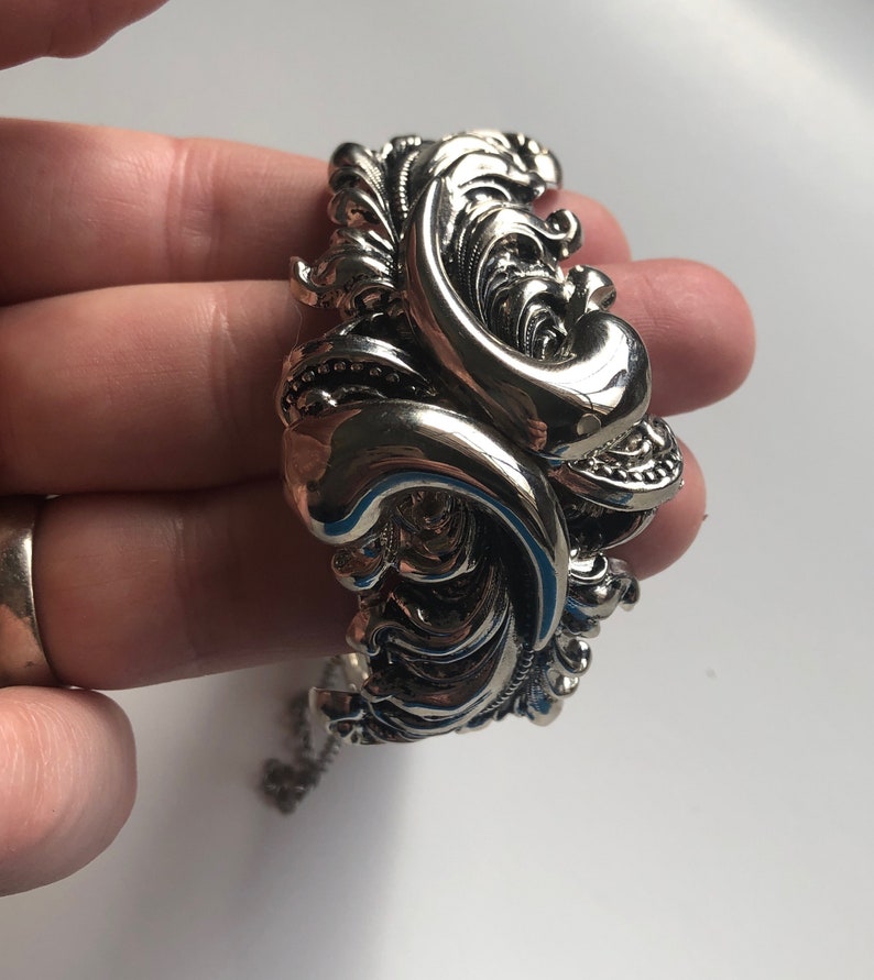 K&ouml;nnte beinhalten: Ein silbernes Armband mit einem verzierten, wirbelnden Design. Das Armband ist offen und hat einen Kettenverschluss.