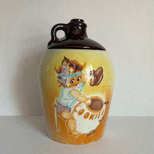 Può includere: Un barattolo per biscotti in ceramica vintage a forma di brocca marrone con un'illustrazione di un bambino con una fascia per capelli che tiene un barattolo per biscotti che dice "Cookies".