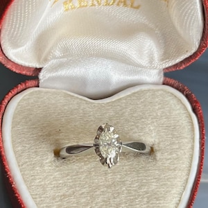 Puede incluir: Un anillo de compromiso de oro blanco con un solo diamante redondo engastado en un halo en forma de coraz&oacute;n. El anillo se presenta en una caja de terciopelo rojo en forma de coraz&oacute;n con la inscripci&oacute;n "H. Hogg Branthewaite Brown Kendal".
