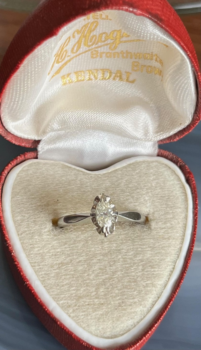 Puede incluir: Un anillo de compromiso de oro blanco con un solo diamante redondo engastado en un halo en forma de coraz&oacute;n. El anillo se presenta en una caja de terciopelo rojo en forma de coraz&oacute;n con la inscripci&oacute;n "H. Hogg Branthewaite Brown Kendal".