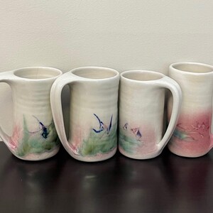 May include: Four white ceramic mugs with large handles. Each mug features a unique watercolour design with pink, green, and blue accents. The mugs are arranged on a dark surface against a neutral background.