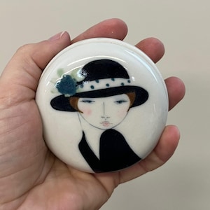 Peut inclure: Une pièce de céramique ronde et blanche représentant une illustration stylisée d'une femme portant un chapeau noir à pois blancs et un ornement floral bleu. La femme a les cheveux bruns, les joues roses et une tenue noire. La pièce est tenue dans une main.