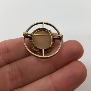May include: A gold and silver toned cameo brooch with a light pink cameo set in a gold toned frame. The brooch is in the shape of a circle and has a pin on the back.
