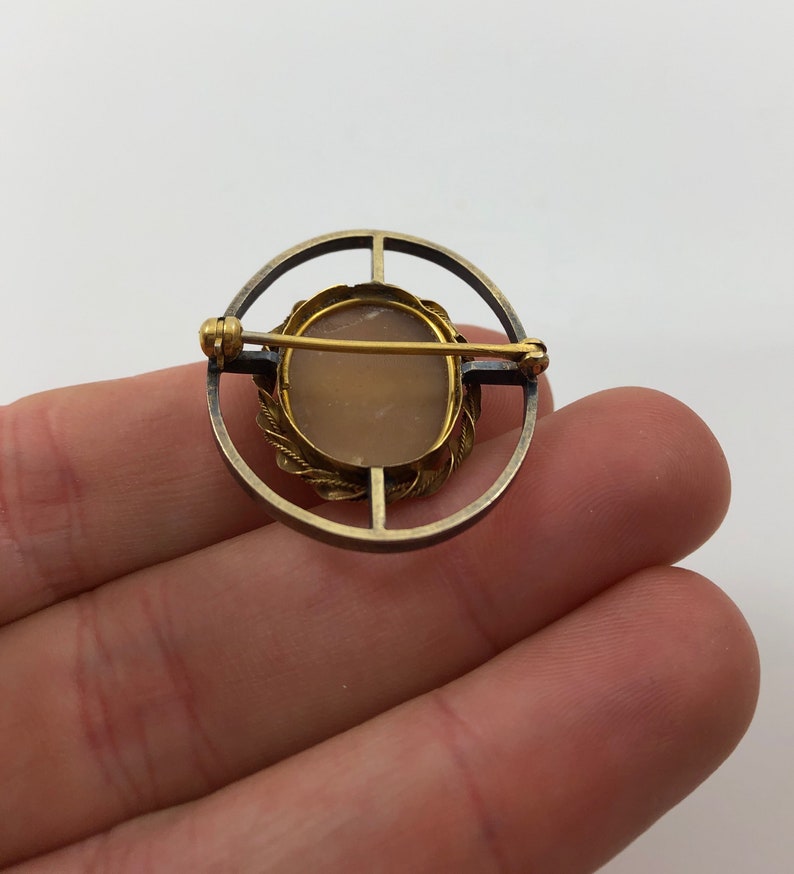 May include: A gold and silver toned cameo brooch with a light pink cameo set in a gold toned frame. The brooch is in the shape of a circle and has a pin on the back.