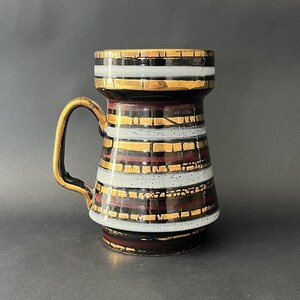 Pode incluir: Uma caneca de cerâmica vintage com uma alça dourada e um corpo cilíndrico. A caneca apresenta faixas horizontais de ouro, branco e marrom escuro, criando um padrão geométrico. As bordas superior e inferior também são acentuadas com ouro.