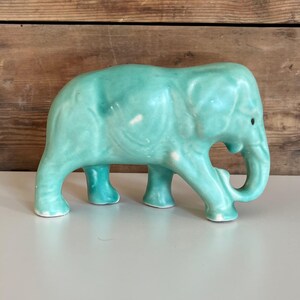 May include: A turquoise ceramic elephant ornament. The elephant has a trunk, tusks, and four legs. The ornament is a solid colour with some variations in the glaze. The elephant is standing on a white surface.