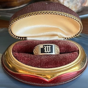 May include: A gold ring with a black rectangular centre displaying the letter "M" in white. The ring is presented in a heart-shaped jewellery box with a red velvet lining and gold trim.