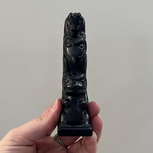 Könnte beinhalten: Kleine, schwarze Totempfahl-Schnitzerei mit drei vertikal gestapelten Figuren. Die oberste Figur ist ein Frosch, die mittlere Figur eine Eule und die untere Figur ein Bär. Der Totempfahl ist etwa 13 cm hoch.