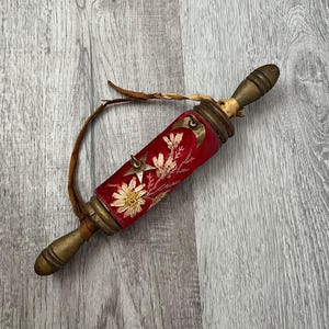 May include: A decorative scroll case with a red velvet exterior, adorned with gold embroidery of flowers and stars. The case has brass accents and wooden handles, with a leather strap attached. The scroll case is approximately 10 inches long.