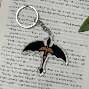 May include: A black and gold dragon keychain with a clear acrylic background. The keychain is hanging from a book with text about a village being attacked. The text reads "Sumerton was attacked."