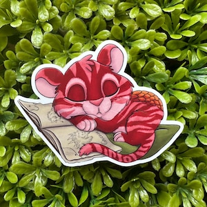 May include: A sticker featuring a cartoon tiger cub with red and pink stripes, sleeping peacefully on a book. The sticker has a white border and is set against a backdrop of green foliage.