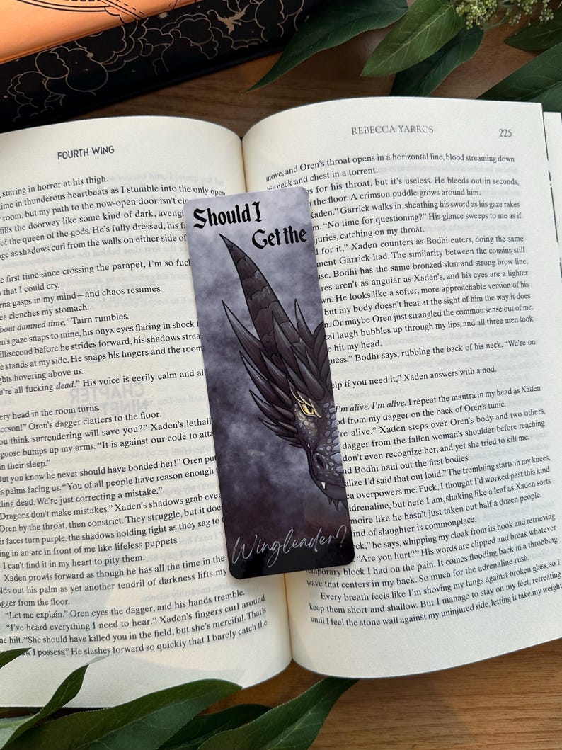 Op de afbeelding: Een close-up van een boek met een bladwijzer met een zwarte en grijze draak met rode ogen. De bladwijzer heeft de tekst "Should I Get the Wingleader?" erop gedrukt.