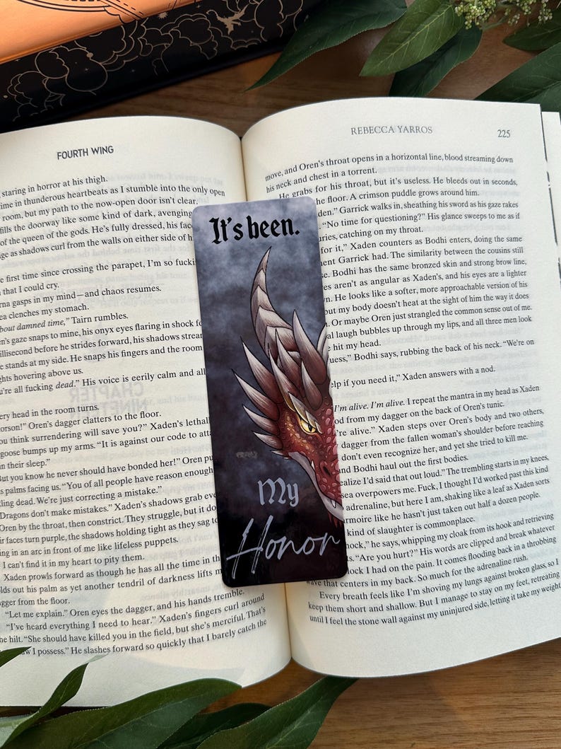 May include: A close-up of a book with a bookmark featuring a red dragon with a black background. The bookmark has the words "My Honor" written in white. The book is open to a page with the text "It's been." The text on the page is about a character named Oren who is bleeding out after being attacked.