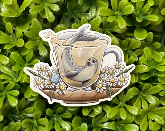 May include: A sticker of a seal swimming in a cup of tea with a tea bag hanging from the rim. The cup is surrounded by white daisies.