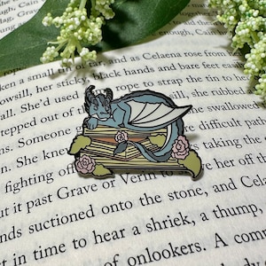 May include: A blue enamel pin of a dragon sleeping on a stack of books with pink flowers. The pin is on a book page with text that reads "She'd used rope to strap the tin to her sill, she rubbed a..."