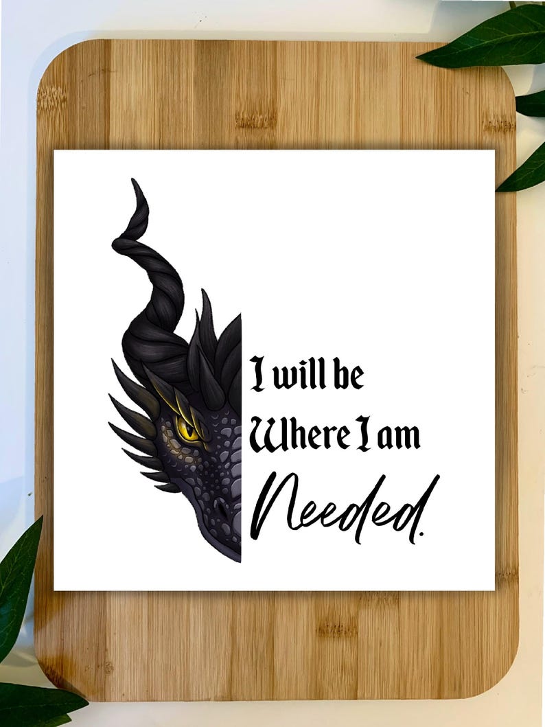 May include: A white square print featuring a dragon's head with a black horn and yellow eye. The text "I will be Where I am Needed" is written in black script. The print is on a wooden cutting board.