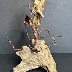 May include: A unique sculpture crafted from driftwood, featuring a stylized face and copper wire accents. The sculpture has a natural, earthy color palette with a textured surface. The artwork is mounted on a piece of driftwood.