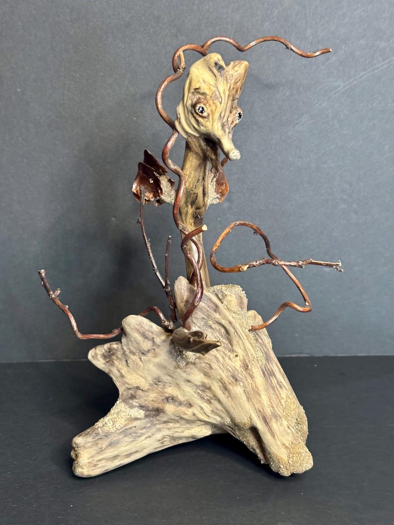May include: A unique sculpture crafted from driftwood, featuring a stylized face and copper wire accents. The sculpture has a natural, earthy color palette with a textured surface. The artwork is mounted on a piece of driftwood.