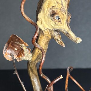 May include: A handcrafted wooden seahorse sculpture. The seahorse is made from natural wood with a light brown color, featuring dark brown vine-like accents. The sculpture has two small, dark eyes and a long snout. The seahorse is mounted on a small wooden base.