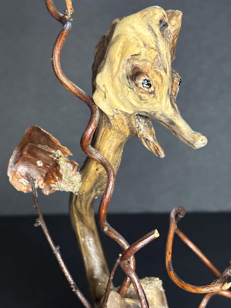 May include: A handcrafted wooden seahorse sculpture. The seahorse is made from natural wood with a light brown color, featuring dark brown vine-like accents. The sculpture has two small, dark eyes and a long snout. The seahorse is mounted on a small wooden base.