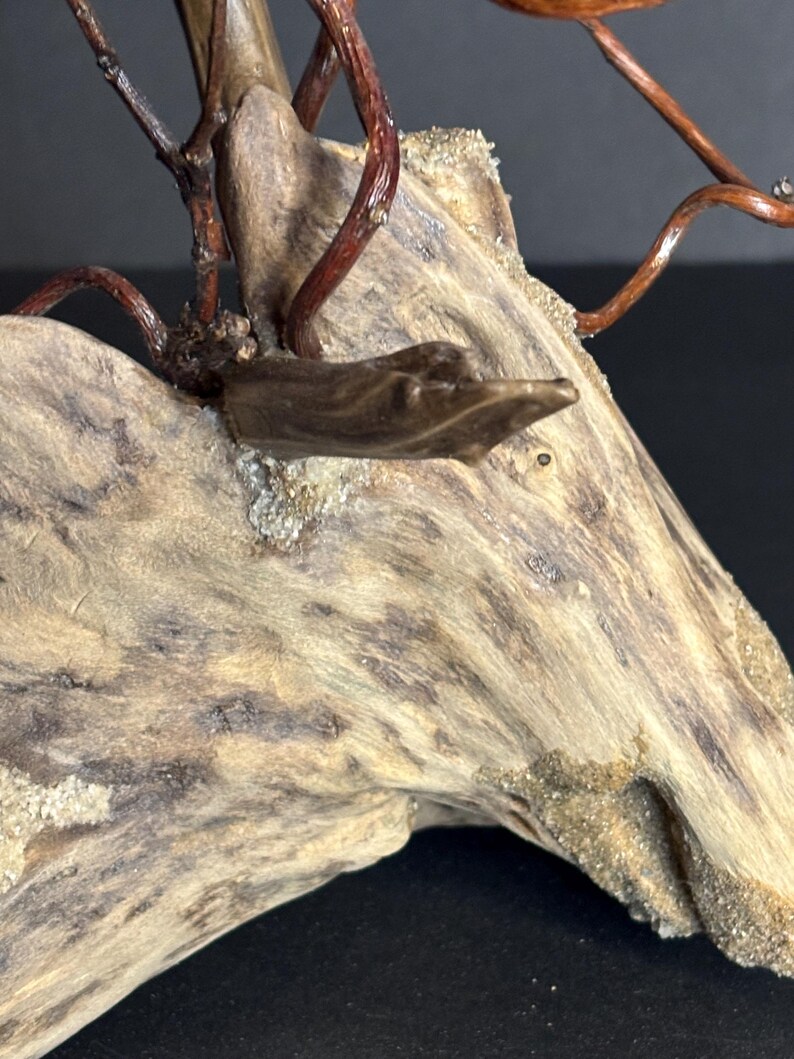 May include: Close-up of a driftwood sculpture with a natural, weathered appearance. The wood is a mix of light and dark browns, with a textured surface. Thin, reddish-brown branches extend from the wood, adding a delicate contrast to the piece.
