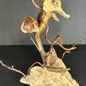 May include: A unique, handcrafted wooden sculpture featuring a whimsical, bird-like figure. The sculpture incorporates natural wood elements, with copper wire accents and small branches. The color palette includes natural wood tones and copper.