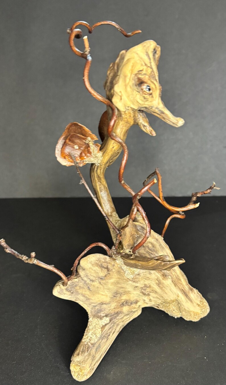 May include: A unique, handcrafted wooden sculpture featuring a whimsical, bird-like figure. The sculpture incorporates natural wood elements, with copper wire accents and small branches. The color palette includes natural wood tones and copper.