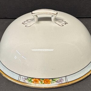 Può includere: Coperchio per piatto da portata in ceramica bianca con manico dorato e bordo floreale. Il coperchio è a forma di cupola con dettagli dorati e una fascia azzurra. Il bordo presenta fiori arancioni e viola.