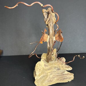 May include: A handcrafted sculpture featuring a central wooden element with a textured, light brown appearance. Copper wire branches extend outward, and amber-colored accents are incorporated. The sculpture rests on a base of light brown, textured wood.