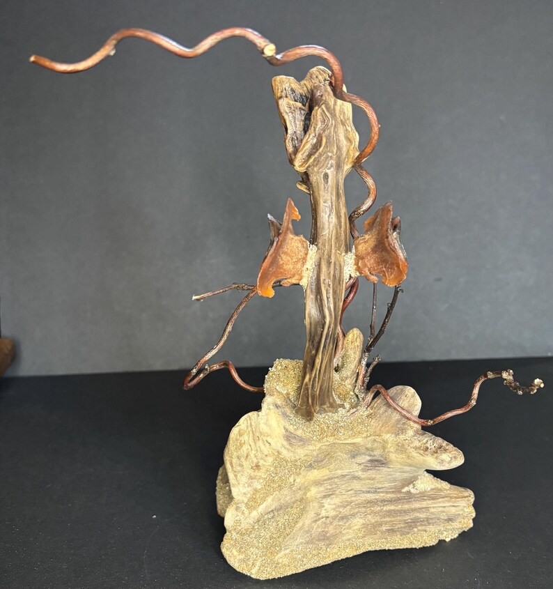 May include: A handcrafted sculpture featuring a central wooden element with a textured, light brown appearance. Copper wire branches extend outward, and amber-colored accents are incorporated. The sculpture rests on a base of light brown, textured wood.