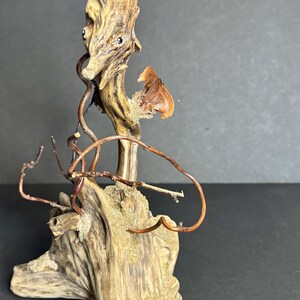 May include: A unique, abstract wooden sculpture with a natural, weathered appearance. The sculpture features a base of driftwood, with copper wire accents and a small, dried leaf. The piece has a neutral color palette with shades of brown and tan.