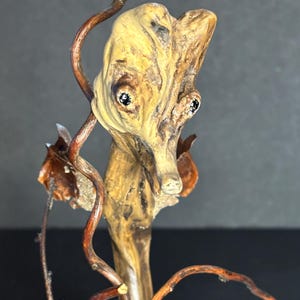 May include: A unique, handcrafted wooden sculpture featuring a stylized face with dark eyes and a long nose. The sculpture is made from natural wood with a light brown color and is accented with dark brown vines.