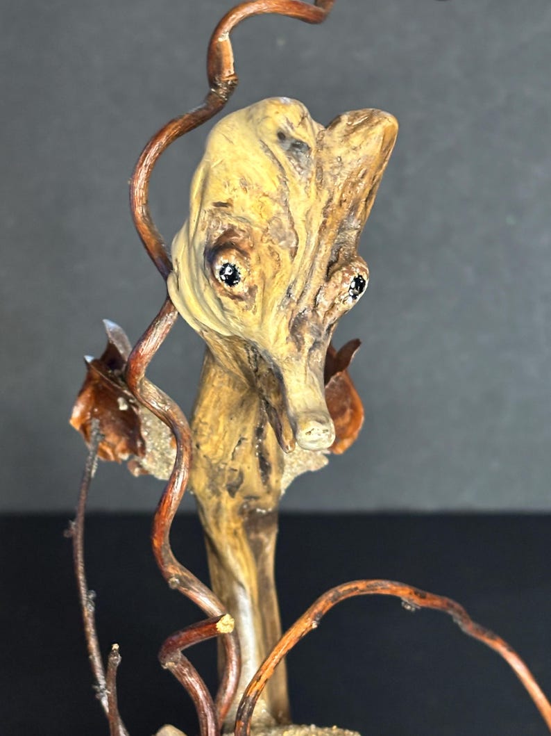 May include: A unique, handcrafted wooden sculpture featuring a stylized face with dark eyes and a long nose. The sculpture is made from natural wood with a light brown color and is accented with dark brown vines.