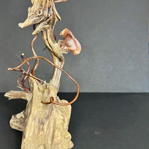 May include: A unique driftwood sculpture featuring a seahorse-like figure with a long snout and curved copper wire accents. The sculpture has a natural wood grain finish with sand-like texture on the base. The overall color palette is brown and copper.