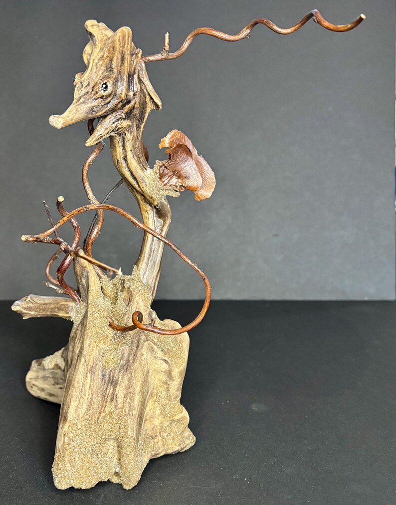 May include: A unique driftwood sculpture featuring a seahorse-like figure with a long snout and curved copper wire accents. The sculpture has a natural wood grain finish with sand-like texture on the base. The overall color palette is brown and copper.