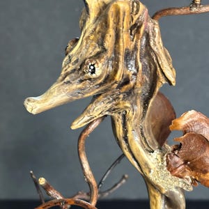May include: A handcrafted wooden seahorse sculpture. The sculpture is made from natural wood, featuring a detailed head, eye, and snout. The seahorse is a light brown color with darker brown accents, and is mounted on a base of twigs and branches.