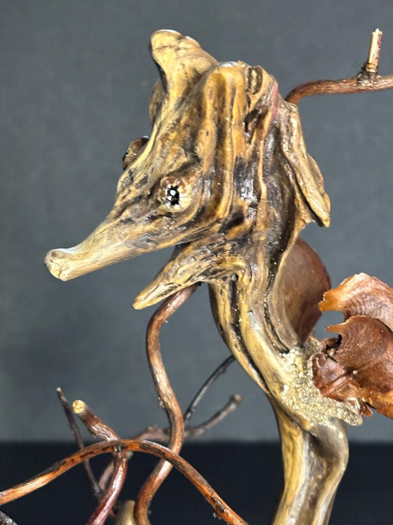 May include: A handcrafted wooden seahorse sculpture. The sculpture is made from natural wood, featuring a detailed head, eye, and snout. The seahorse is a light brown color with darker brown accents, and is mounted on a base of twigs and branches.