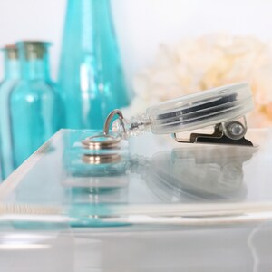 May include: A clear plastic retractable badge holder with a silver clip and a silver ring. The holder is on a clear acrylic surface.