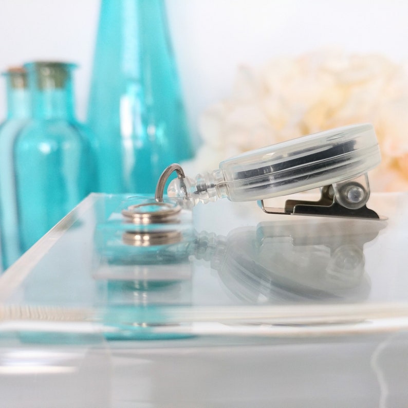 May include: A clear plastic retractable badge holder with a silver clip and a silver ring. The holder is on a clear acrylic surface.