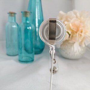 May include: A white retractable badge holder with a clear plastic reel and a silver clip. The holder is on a white stand.