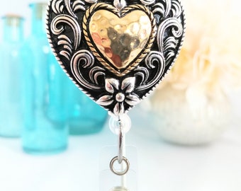 May include: A silver and gold heart-shaped badge holder with a decorative floral design. The heart has a hammered gold finish and a black rope border.