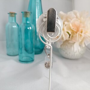 May include: White retractable badge holder with a clear plastic badge holder and a silver clip. The holder is attached to a white stand.