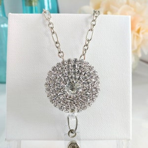 May include: A silver chain with a large, round rhinestone pendant. The pendant is made of many small rhinestones arranged in a circular pattern. The chain has a clear plastic badge holder attached to it.