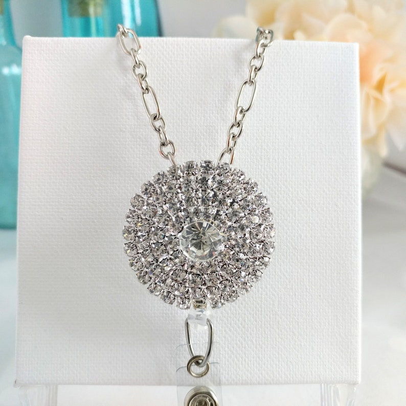 May include: A silver chain with a large, round rhinestone pendant. The pendant is made of many small rhinestones arranged in a circular pattern. The chain has a clear plastic badge holder attached to it.