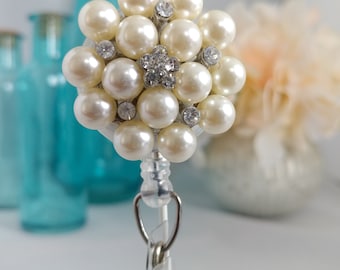 May include: A white pearl and rhinestone flower charm attached to a silver retractable badge holder.