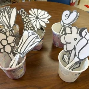 Peut inclure: Plusieurs découpes de fleurs en papier de différents modèles, dont des marguerites et des tulipes, sont disposées dans de petits gobelets blancs. Les fleurs sont en noir et blanc, et les gobelets sont sur une table en bois brun.