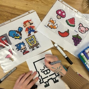 May include: Pixel art templates featuring characters like Mario, Sonic, and SpongeBob, alongside other designs such as a fox, mushroom, and watermelon. A person is drawing a pixelated character with a black marker on graph paper.