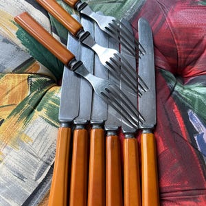 May include: A set of vintage silverware, including forks and knives. The knives have stainless steel blades and orange-brown handles. The forks are stainless steel. The silverware is arranged on a patterned surface.