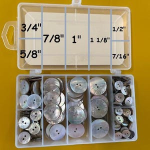 May include: A clear plastic storage box with a variety of white pearl buttons in different sizes. The box has labels indicating the size of each button: 3/4", 7/8", 1", 1 1/8", 1/2", 7/16", and 5/8".