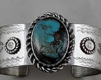May include: A silver cuff bracelet with a turquoise stone set in a rope design. The bracelet is decorated with engraved designs and silver accents.